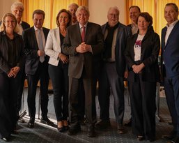 Eine Gruppe von zehn Personen steht in einem Raum, einige lächeln, während sie sich um einen Mann in der Mitte gruppieren, der lässig gekleidet ist. Die anderen sind formell gekleidet und scheinen an einer Besprechung teilzunehmen.