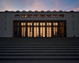Das Gebäude zeigt eine moderne Fassade mit großen Glasfenstern und mehreren vertikalen Säulen. Die Stufen führen zur Eingangstür, die von warmem Licht beleuchtet wird, während der Himmel in sanften Farben erstrahlt.