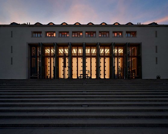 Das Gebäude zeigt eine moderne Fassade mit großen Glasfenstern und mehreren vertikalen Säulen. Die Stufen führen zur Eingangstür, die von warmem Licht beleuchtet wird, während der Himmel in sanften Farben erstrahlt.