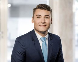 Ein junger Mann in einem dunklen Anzug und hellblauer Krawatte sitzt lächelnd vor einem modernen Hintergrund mit großen Fenstern.