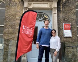 Zwei Personen stehen vor dem Eingang des King's College London, neben einem roten Banner mit dem Logo der Institution. Der Mann trägt einen blauen Pullover und die Frau ein weißes Oberteil.