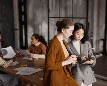 Two women are standing at a table, looking at a tablet together. In the background, several people are participating in a meeting, with documents and cups on the table.